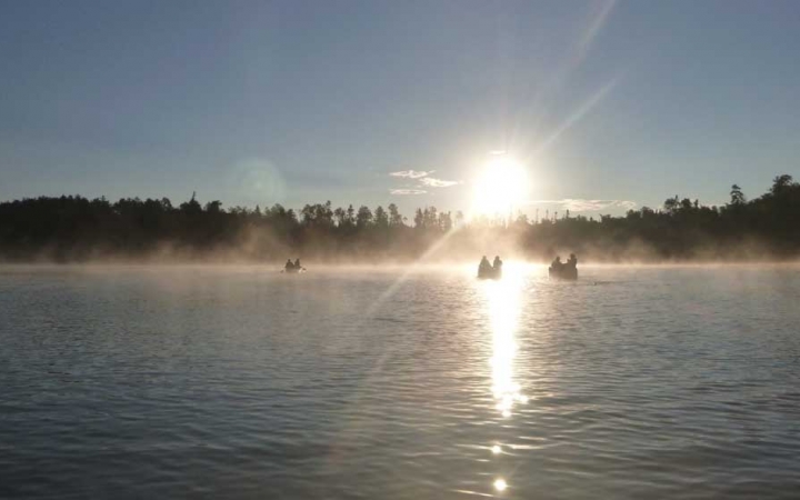 Fog rests on a calm body of water as the sun rises above the tree line in the distance. On the water, three canoes sit.